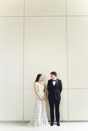 A couple in wedding attire stands side by side against a plain white wall, creating a minimalist elegant composition.