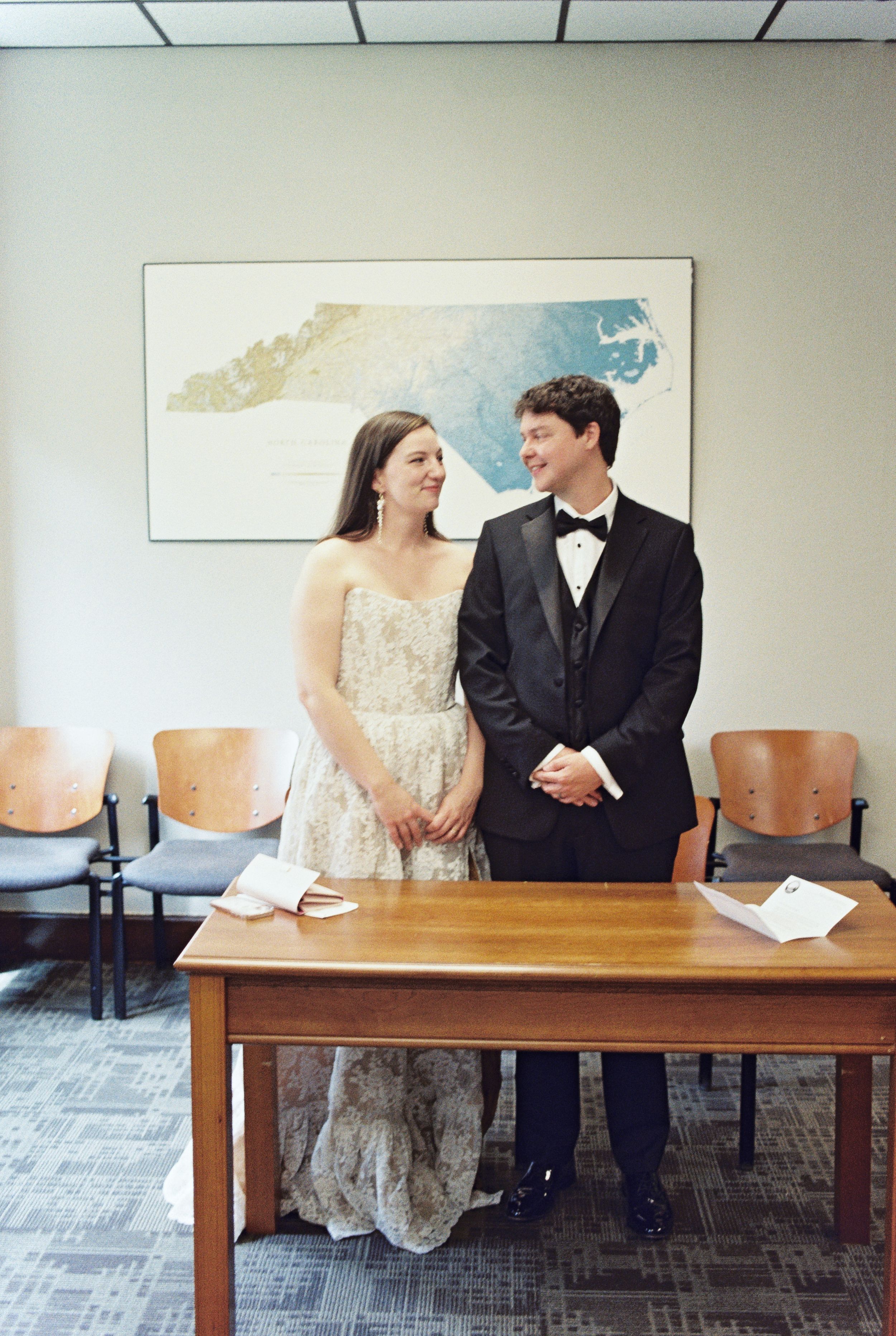 A couple in formal attire, including a white lace dress and black tuxedo, stand together at a wooden table in an office setting.