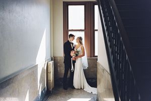 A couple in elegant wedding attire share an intimate moment in a sunlit stairwell hallway.