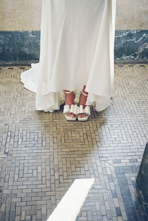 A white wedding dress hem and elegant white platform sandals on a vintage herringbone tile floor.