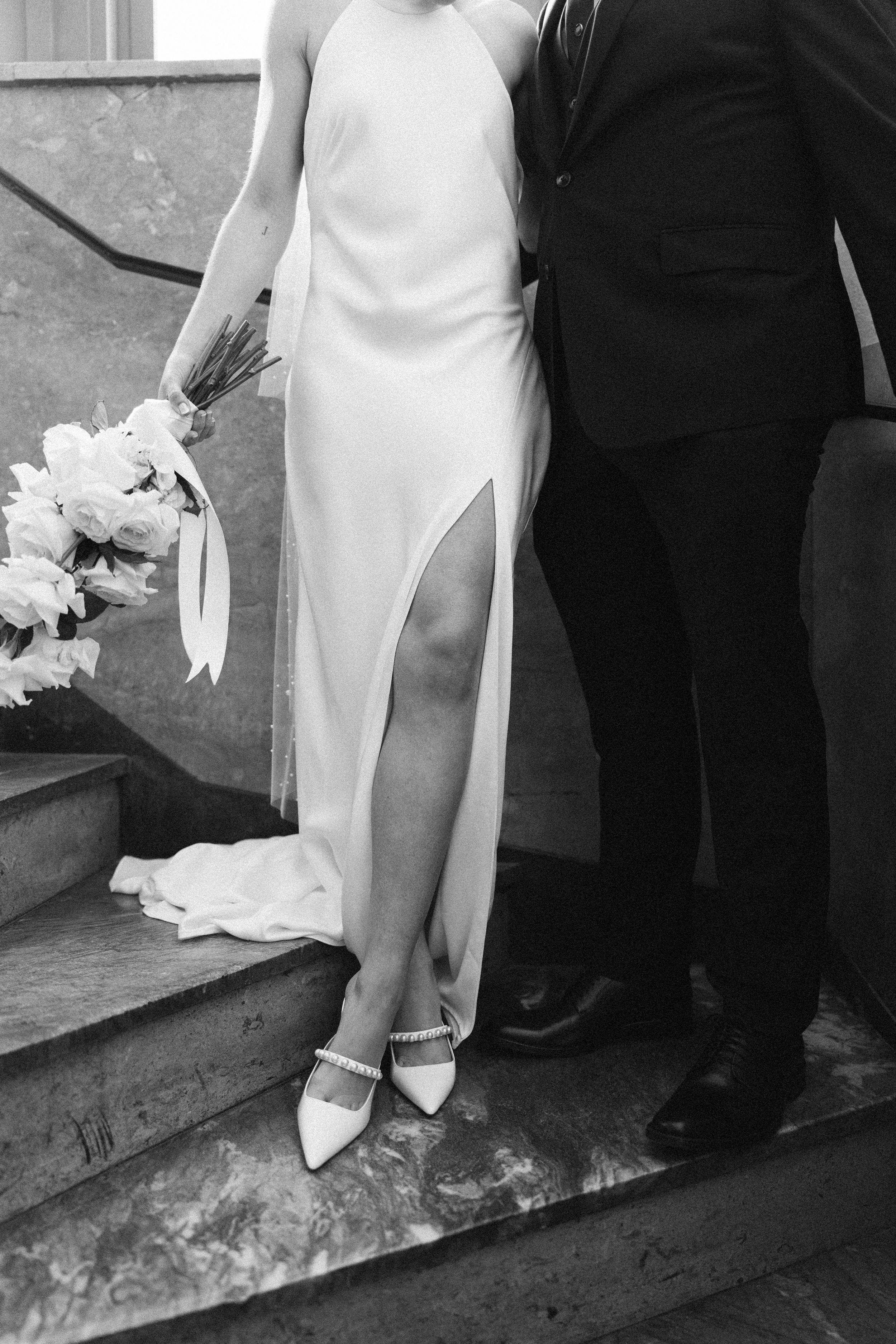 Black and white wedding photo showing bride in elegant white slit dress holding bouquet standing next to groom on steps.