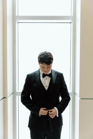 Someone in a classic black tuxedo adjusts their bow tie while standing in a bright white hallway before an event.