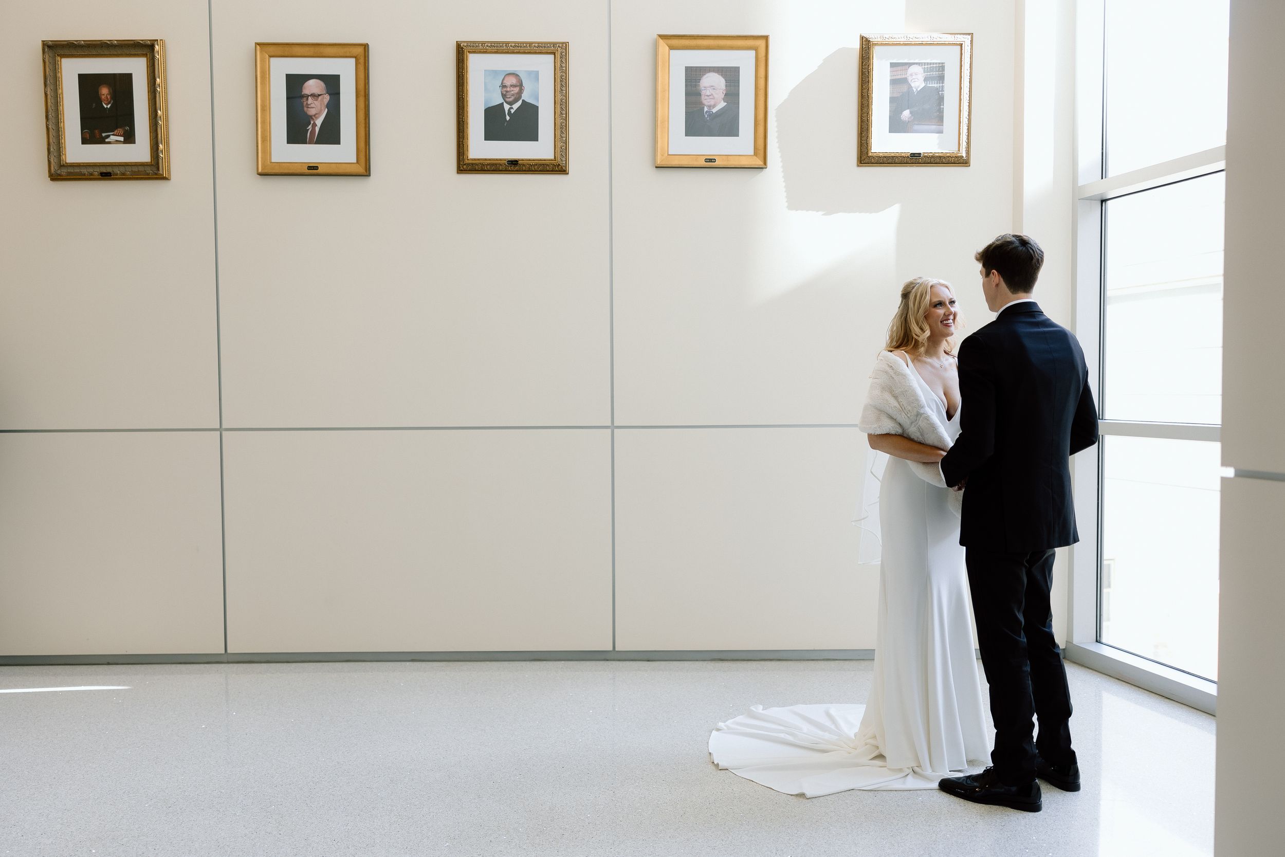 Two people in formal attire view framed photographs displayed on a white gallery wall near a bright window.