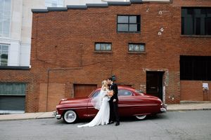 A newlywed couple poses near a classic burgundy car against a rustic brick building backdrop.