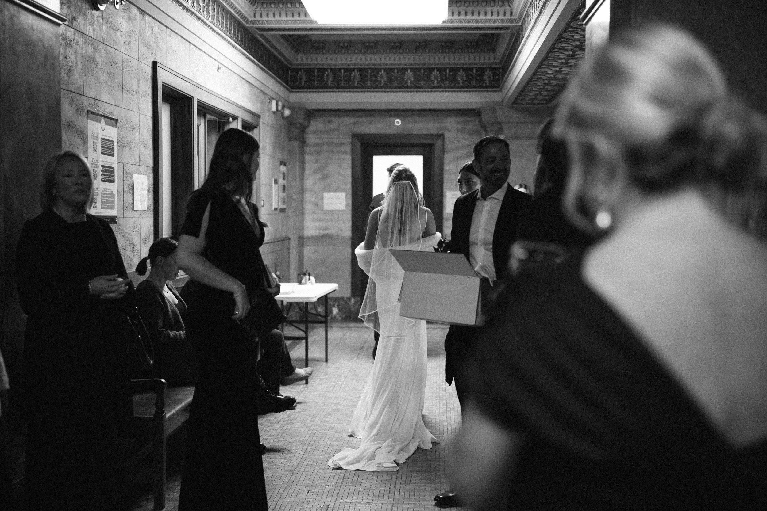 A black and white photograph of people gathering in a formal hallway during what appears to be a wedding event.
