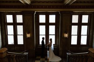 Silhouetted figures stand in dark wooden hall with tall windows and warm wall sconces casting dramatic light.
