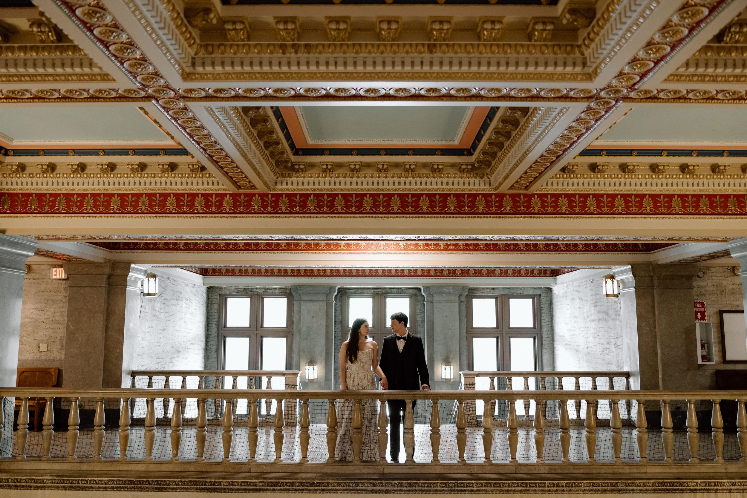 Two figures stand in a luxurious historic venue with ornate gold ceiling details and elegant architectural elements.