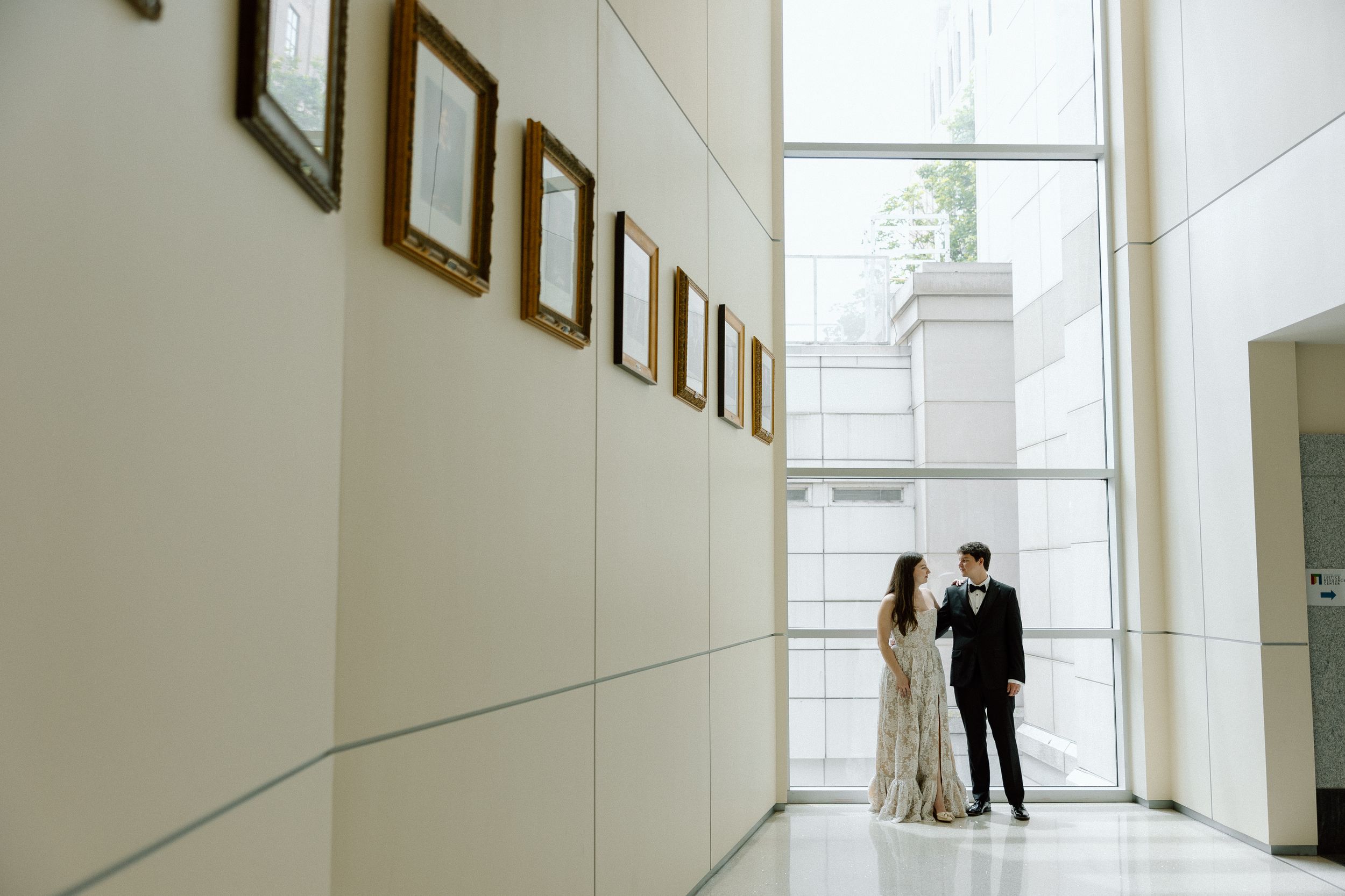 Two dark silhouettes stand in conversation at the end of a minimalist white hallway with large windows.