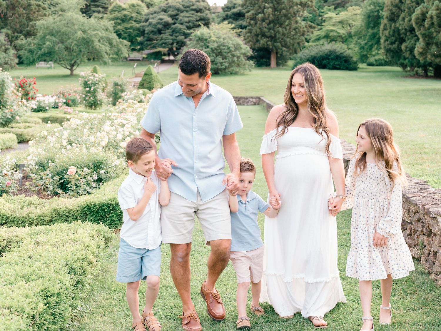 Family of soon to be six holding hands and walking up a stone path surrounded by colorful summer blooms.