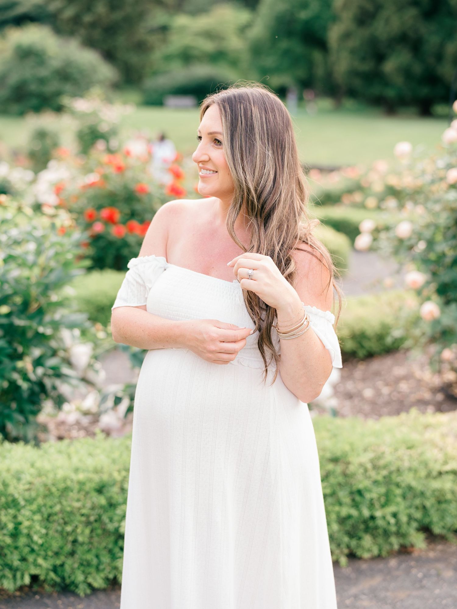 Pregnant woman plays with her long flowing hair standing in profile in front of a blooming rose bush.