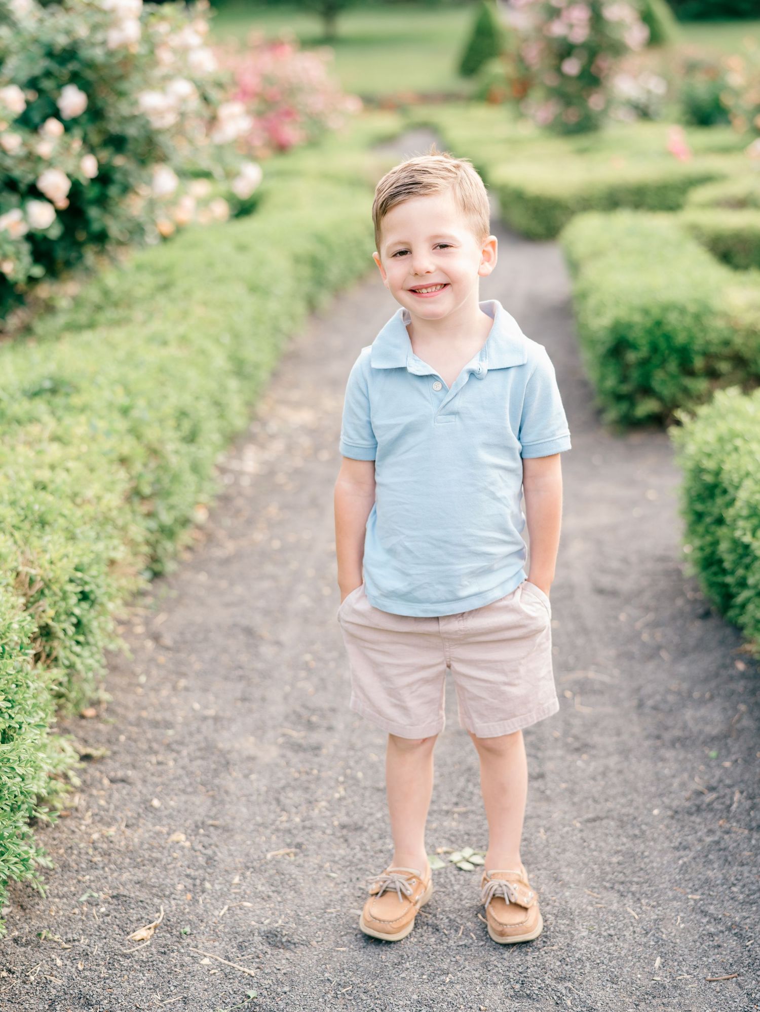 Boy in a blue shirt smiling while standing in a pathway in a garden.