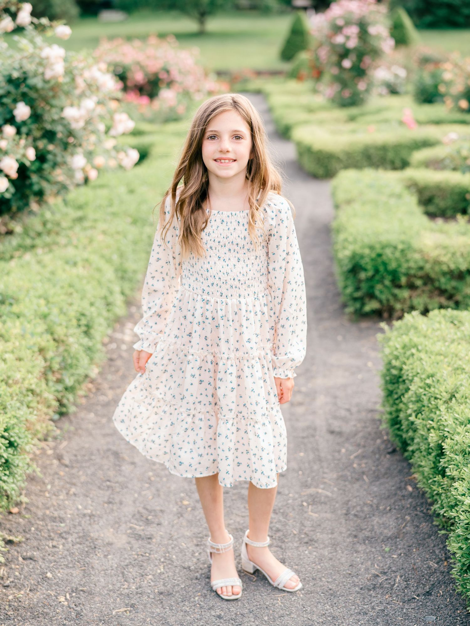 A portrait of a 9 year old girl in a floral dress standing in a garden of roses.