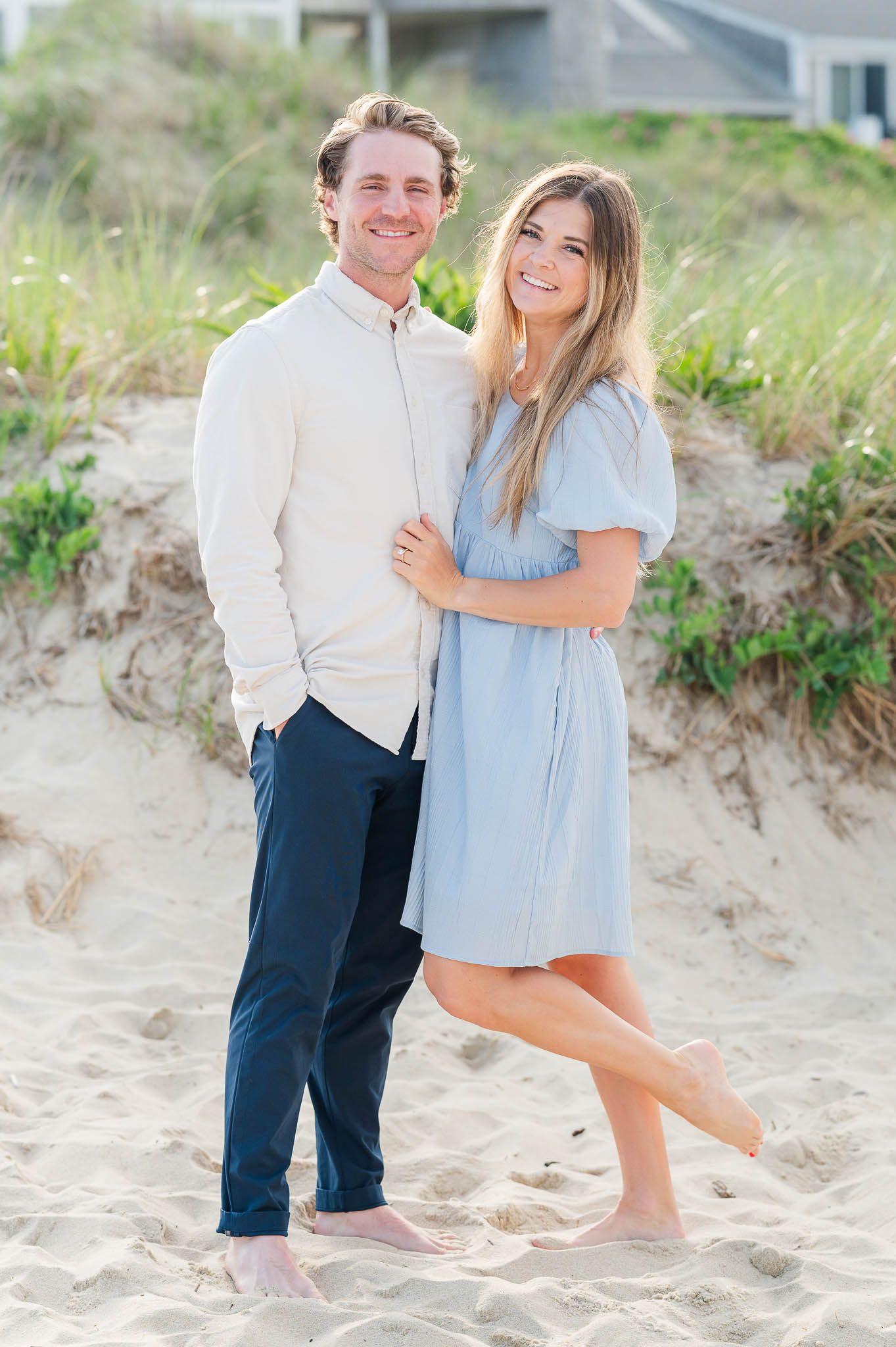 Couple poses together on sandy beach with woman in blue dress and man in white shirt on sunny summer day.