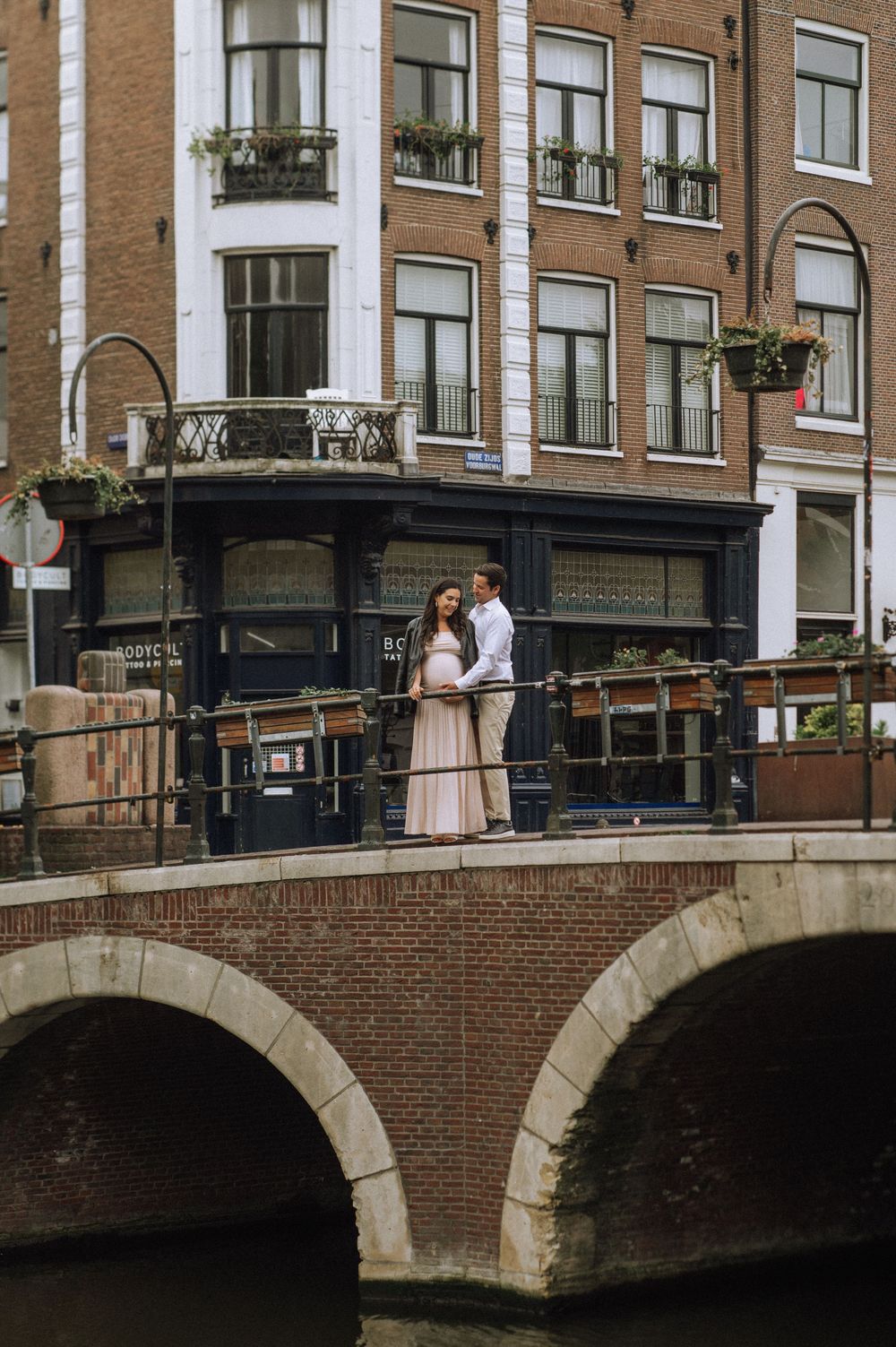 A couple stands together on a brick bridge over an Amsterdam canal with traditional Dutch buildings behind them.