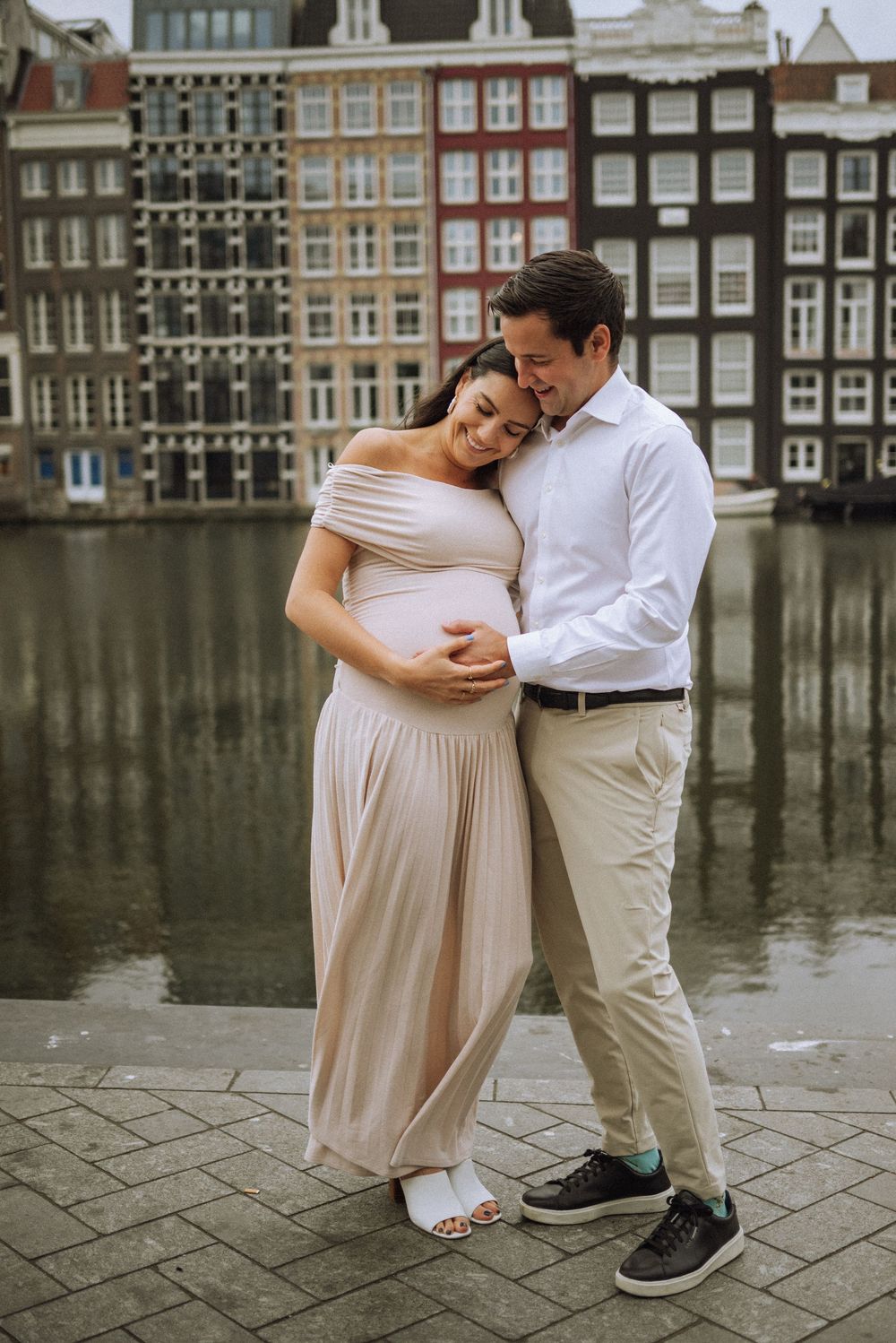 A couple in light-colored clothing poses romantically along Amsterdam's iconic canal waterfront.
