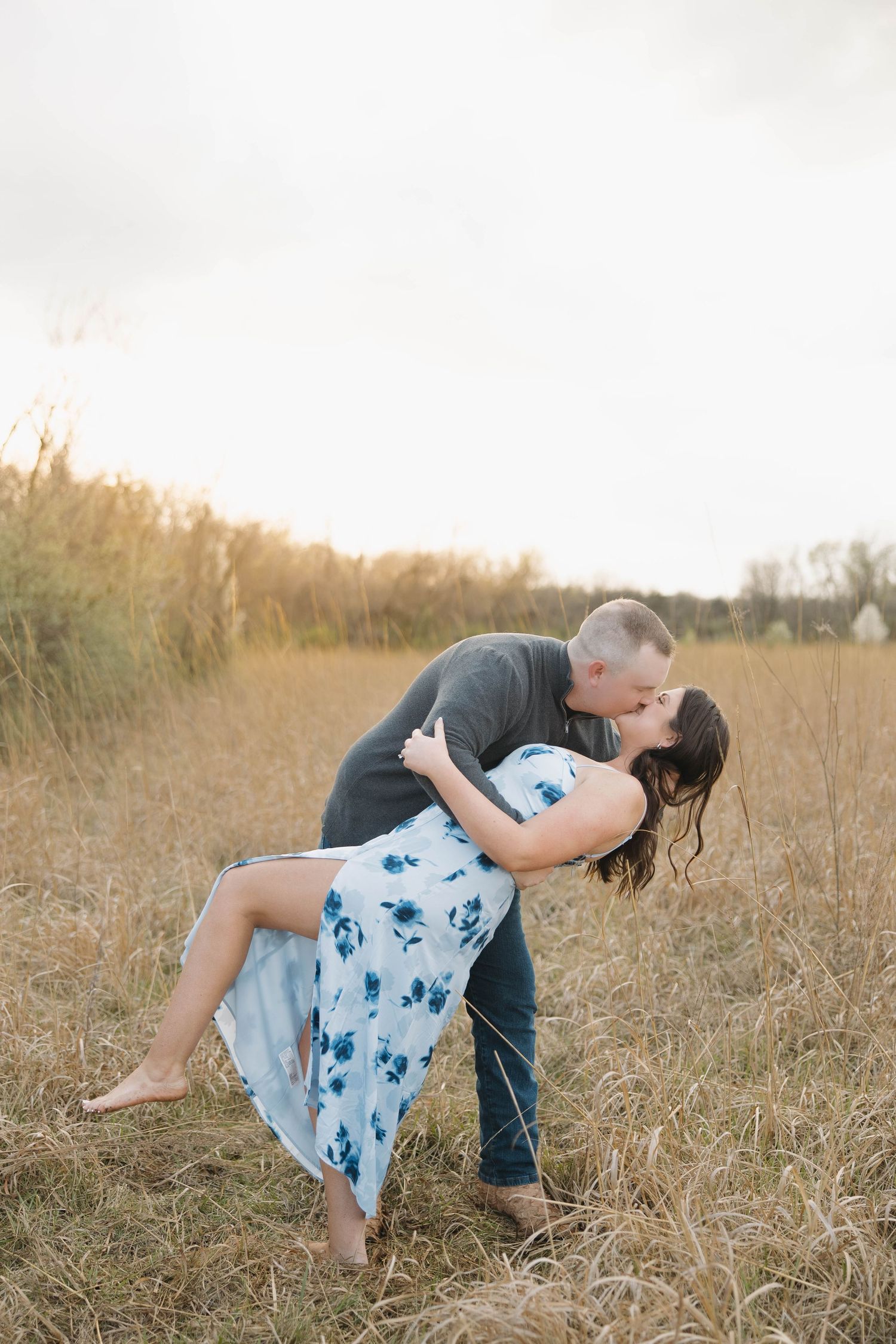 Two people share a romantic moment in a golden field at sunset, with tall grass and trees in the background.