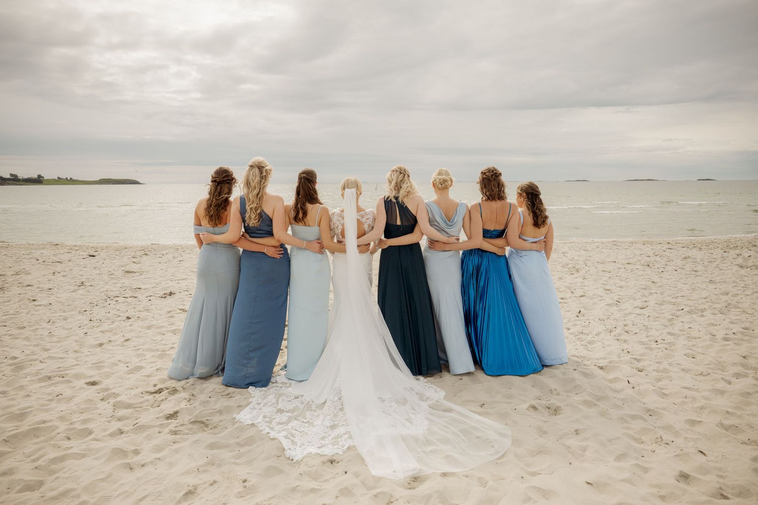 Bridesmaids wearing different shades of blue dresses stand with bride on beach looking at ocean view.