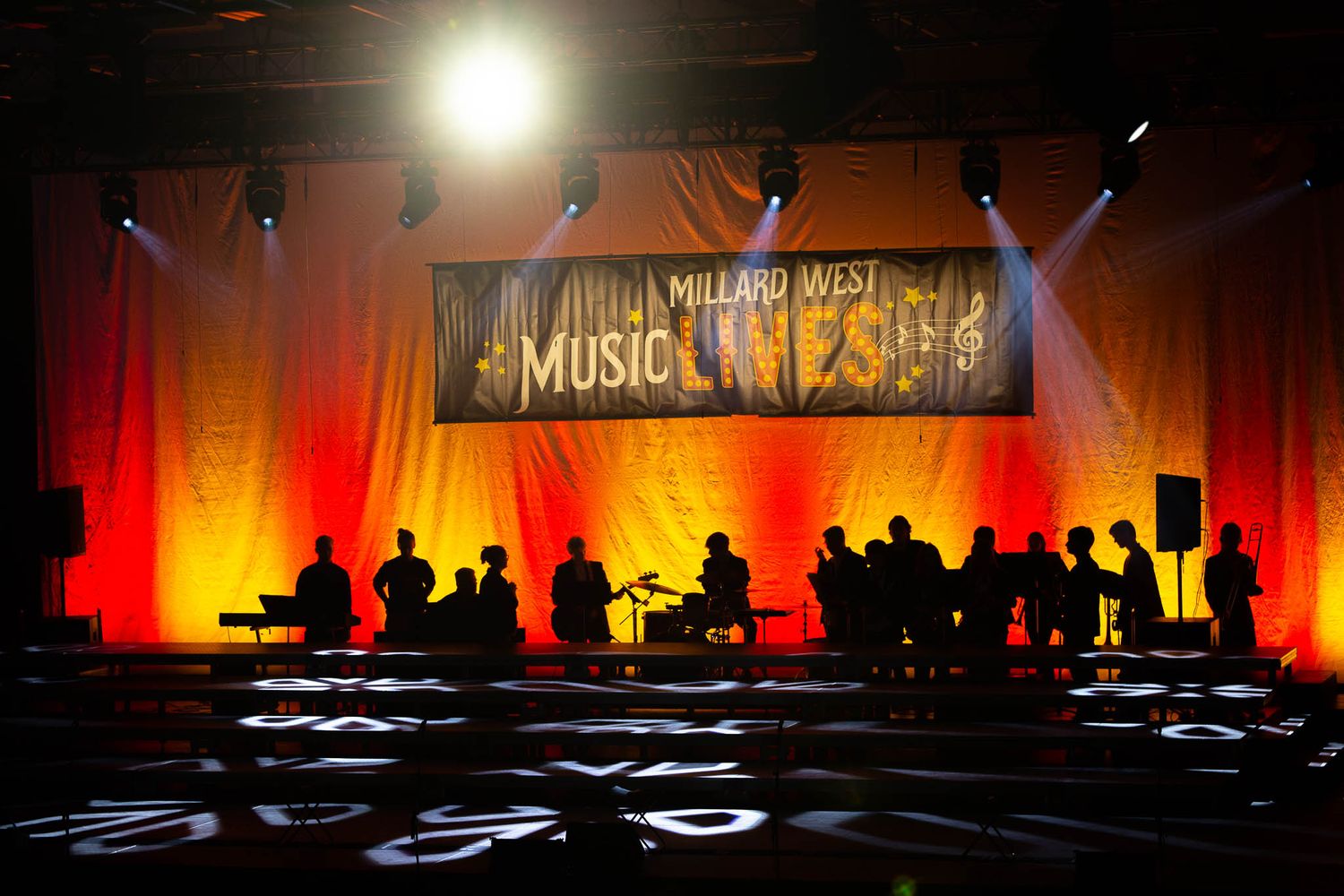 A theatrical stage performance with dramatic spotlights shining down on a group of performers arranged in tiered rows.