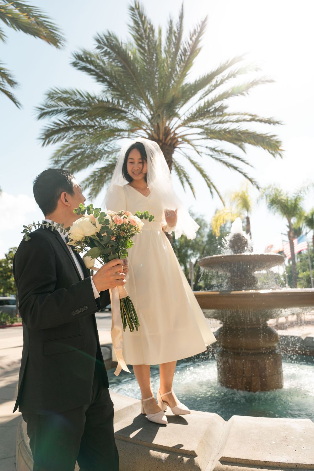 A couple shares a romantic moment by a fountain with palm trees, featuring a white tea-length dress and formal attire.