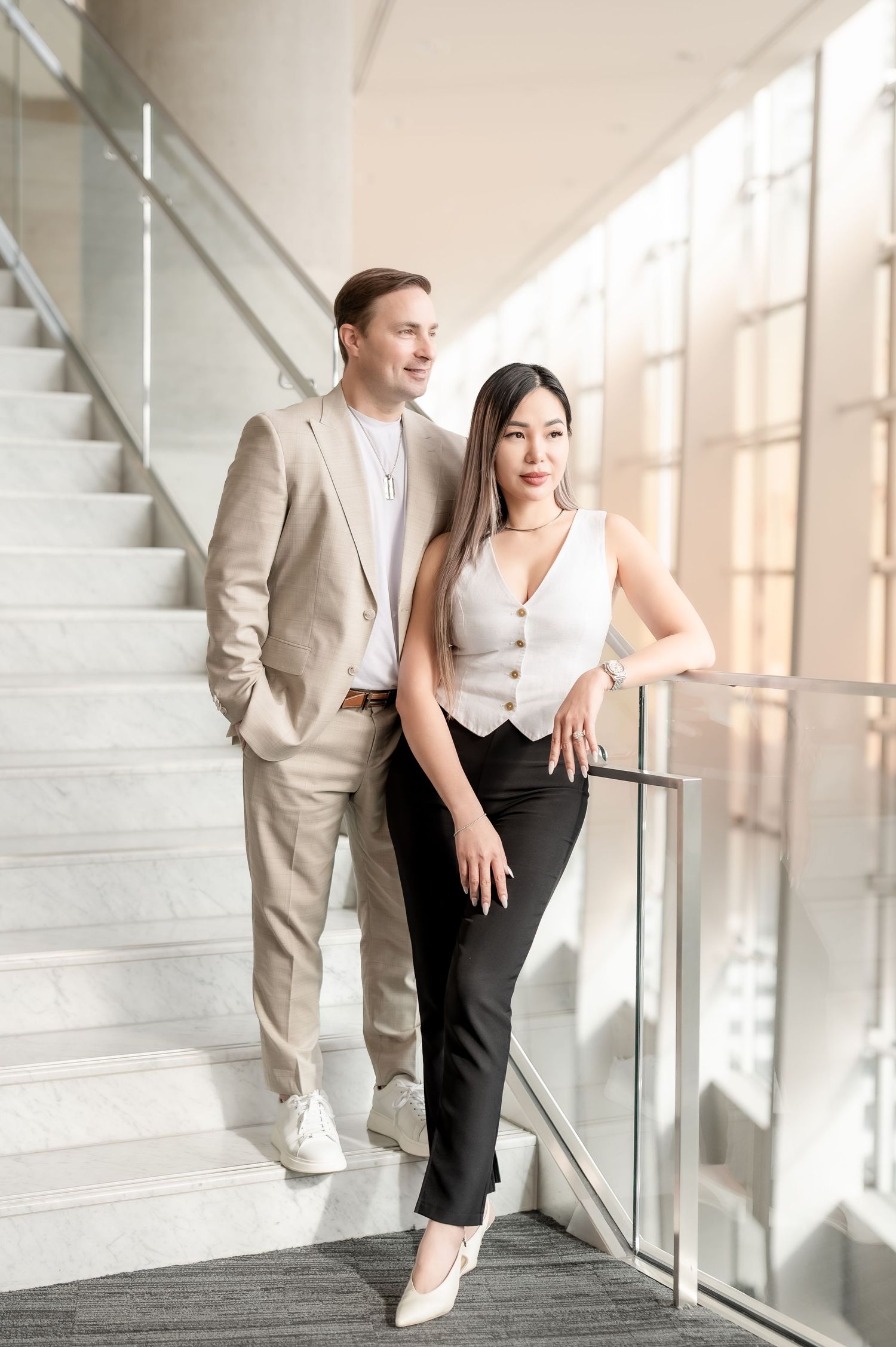 Professional business couple in formal attire poses in modern office building with natural lighting.