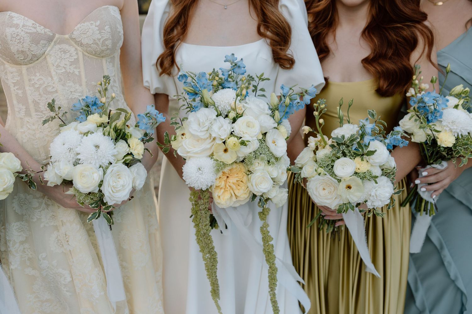 Bridesmaids holding elegant white and cream floral bouquets with blue accents against sage green dresses.