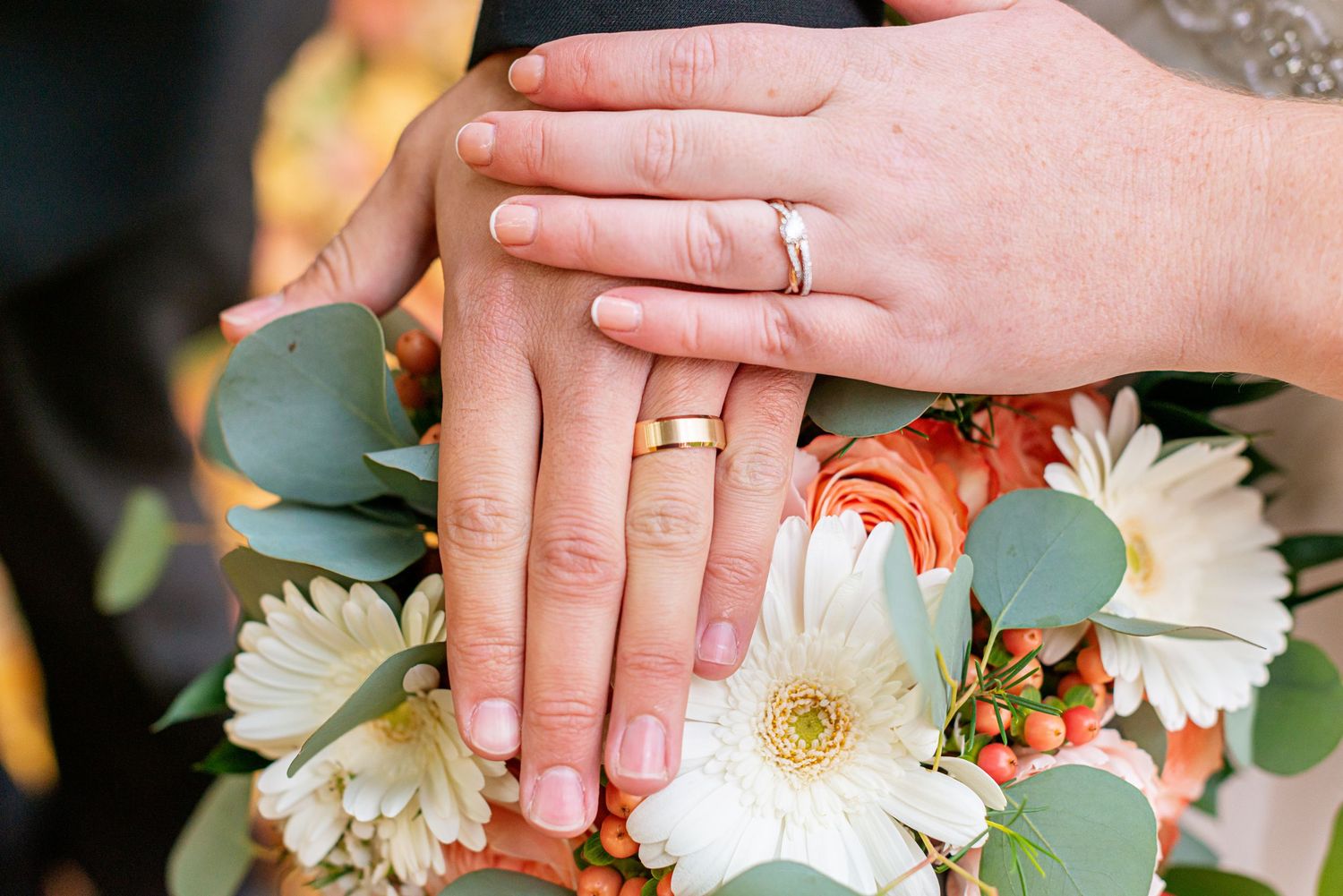 Wedding rings displayed on a bouquet of white daisies and eucalyptus leaves against a dark background.