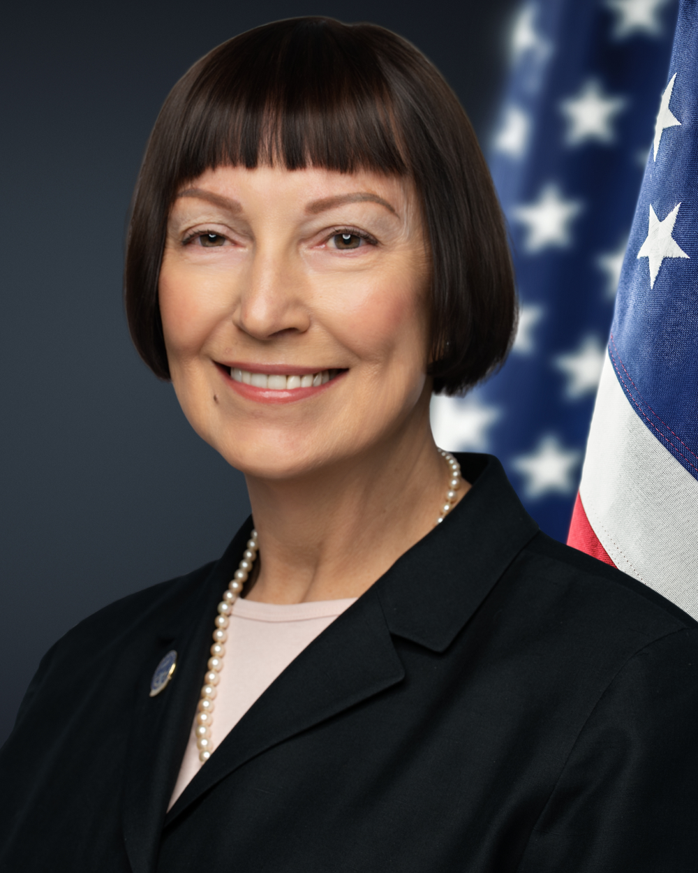 Professional portrait of a person wearing a black blazer and pearl necklace with an American flag backdrop.