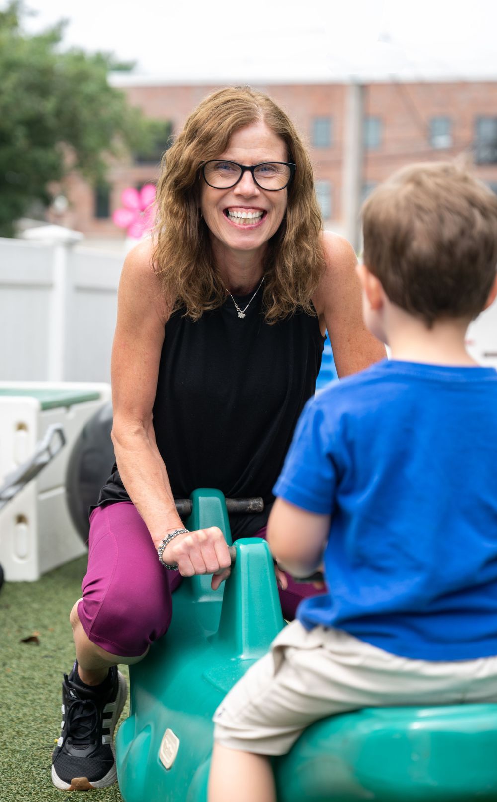 A person wearing a black sleeveless top and purple pants plays with a child on a teal playground seesaw.