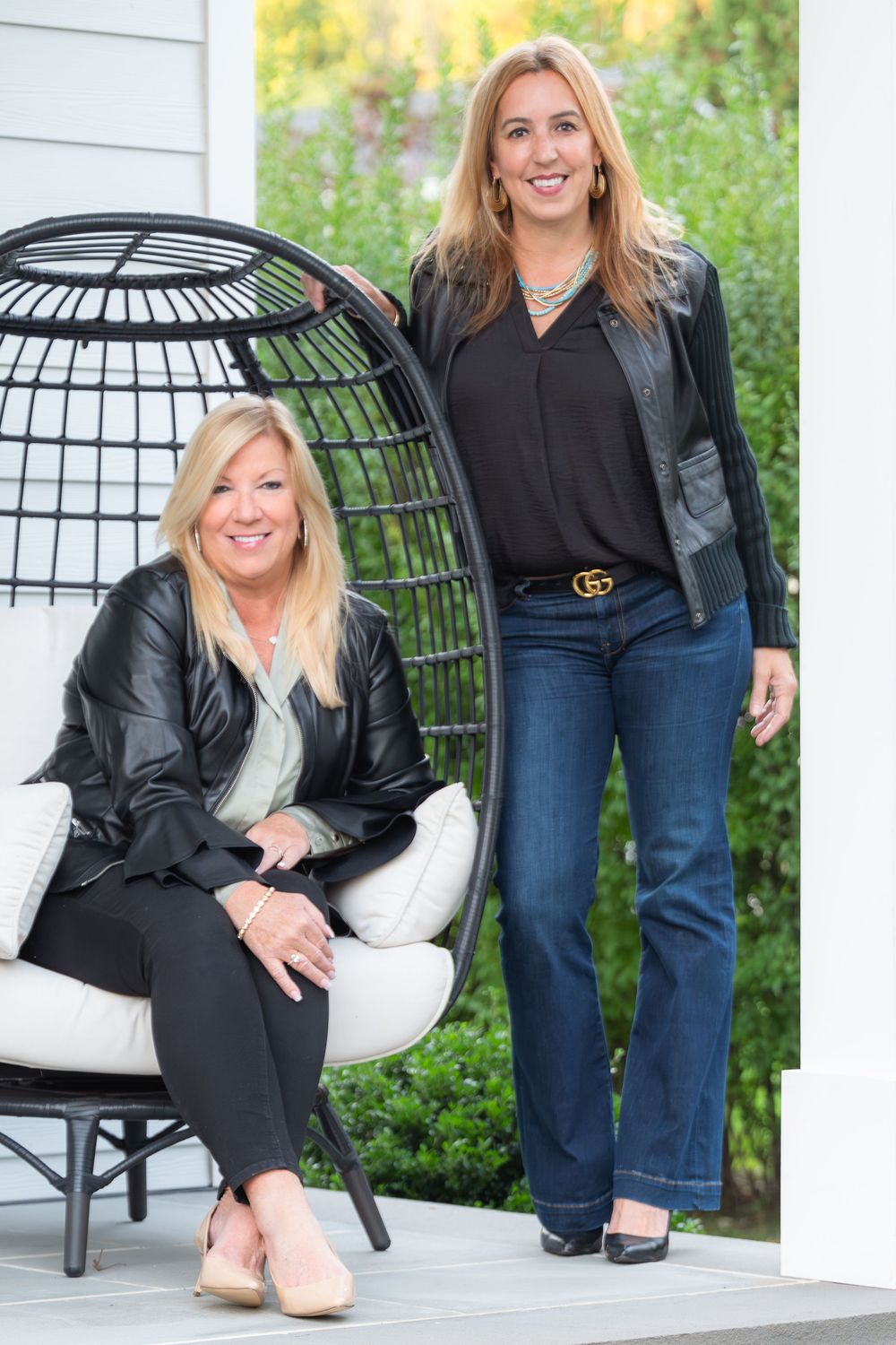 Two women in black outfits pose outdoors, one seated in a black wicker chair and one standing, with greenery in background.