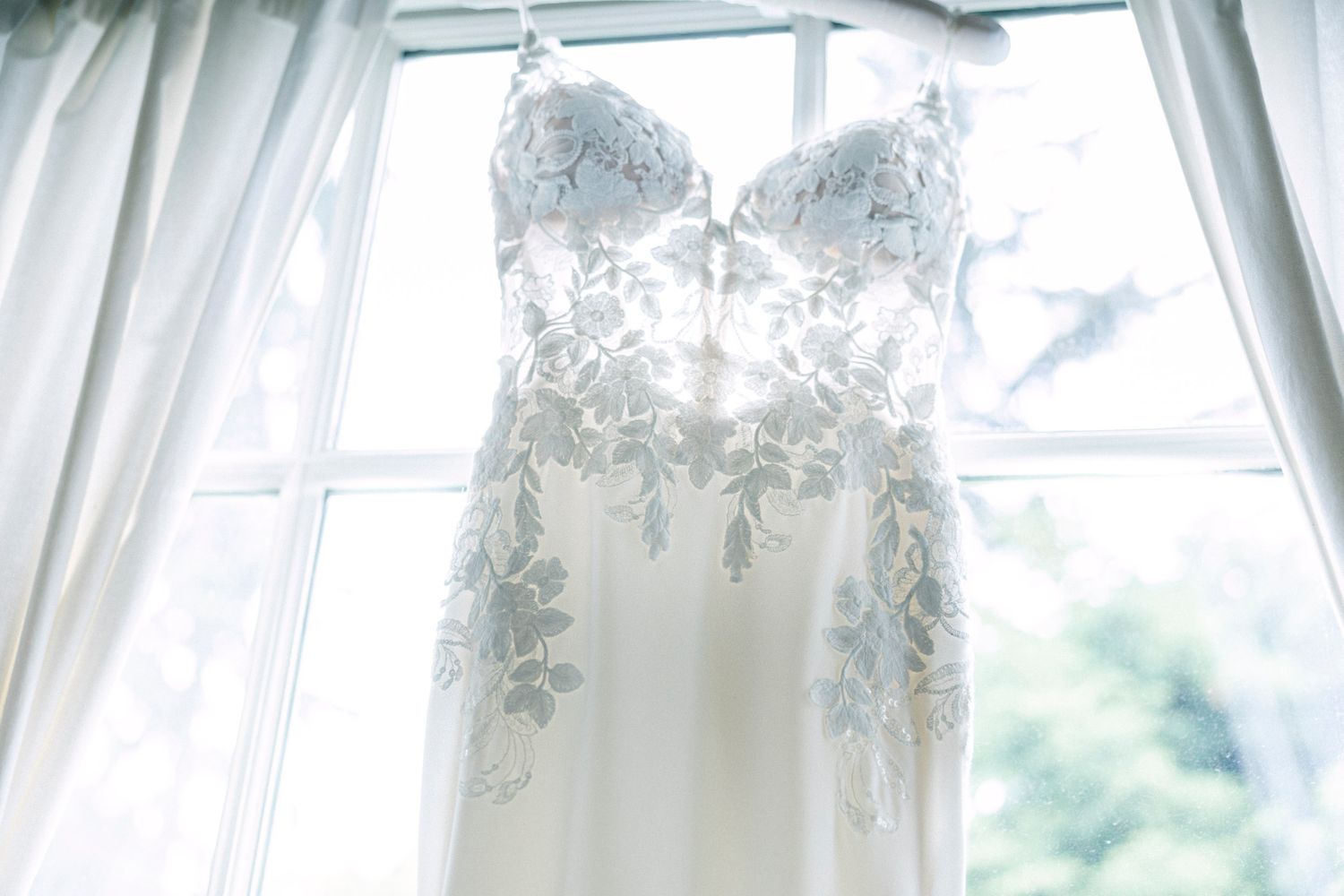 White lace wedding dress hanging in front of sunlit windows with sheer curtains.