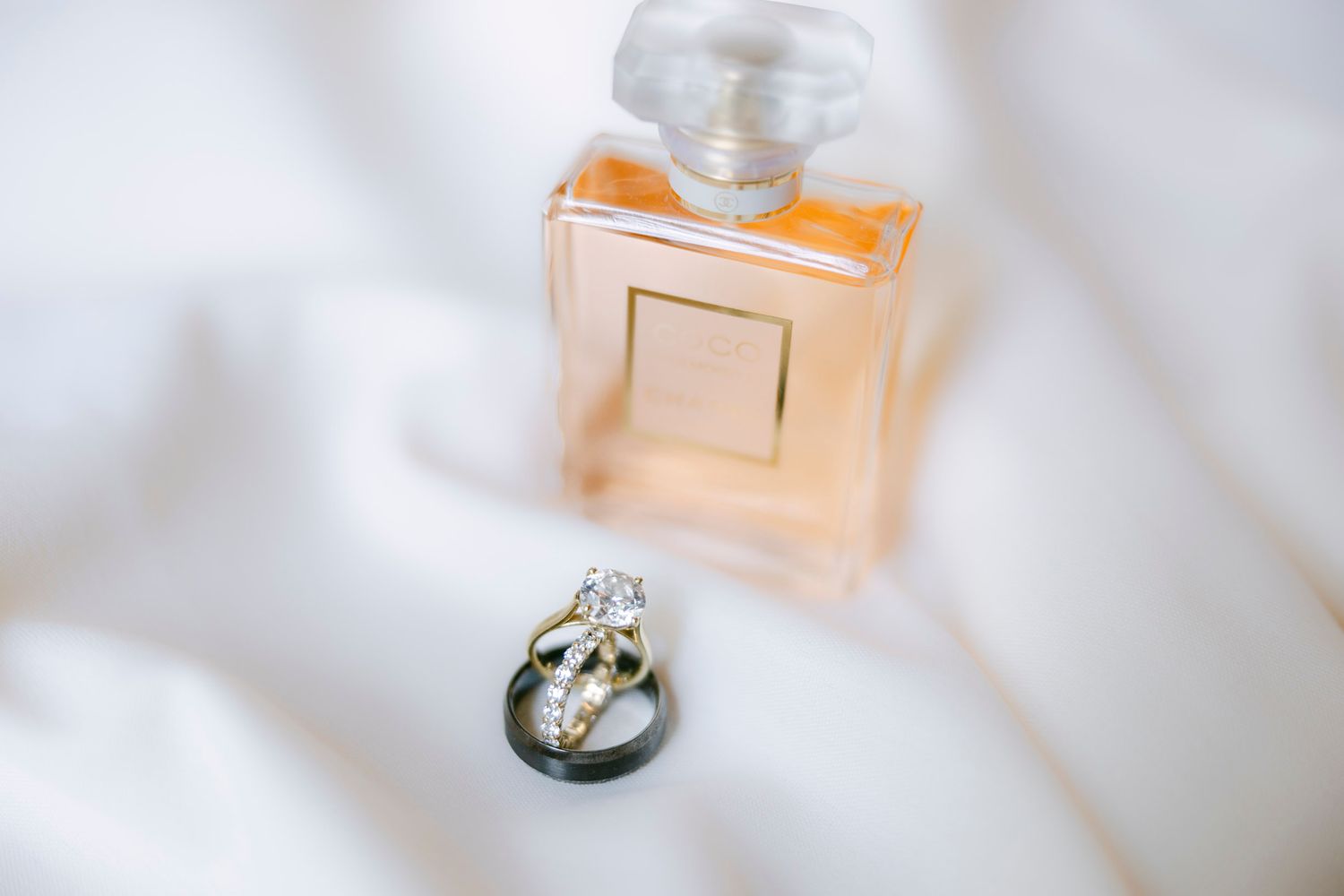 A perfume bottle and wedding rings displayed on white fabric with soft focus lighting.