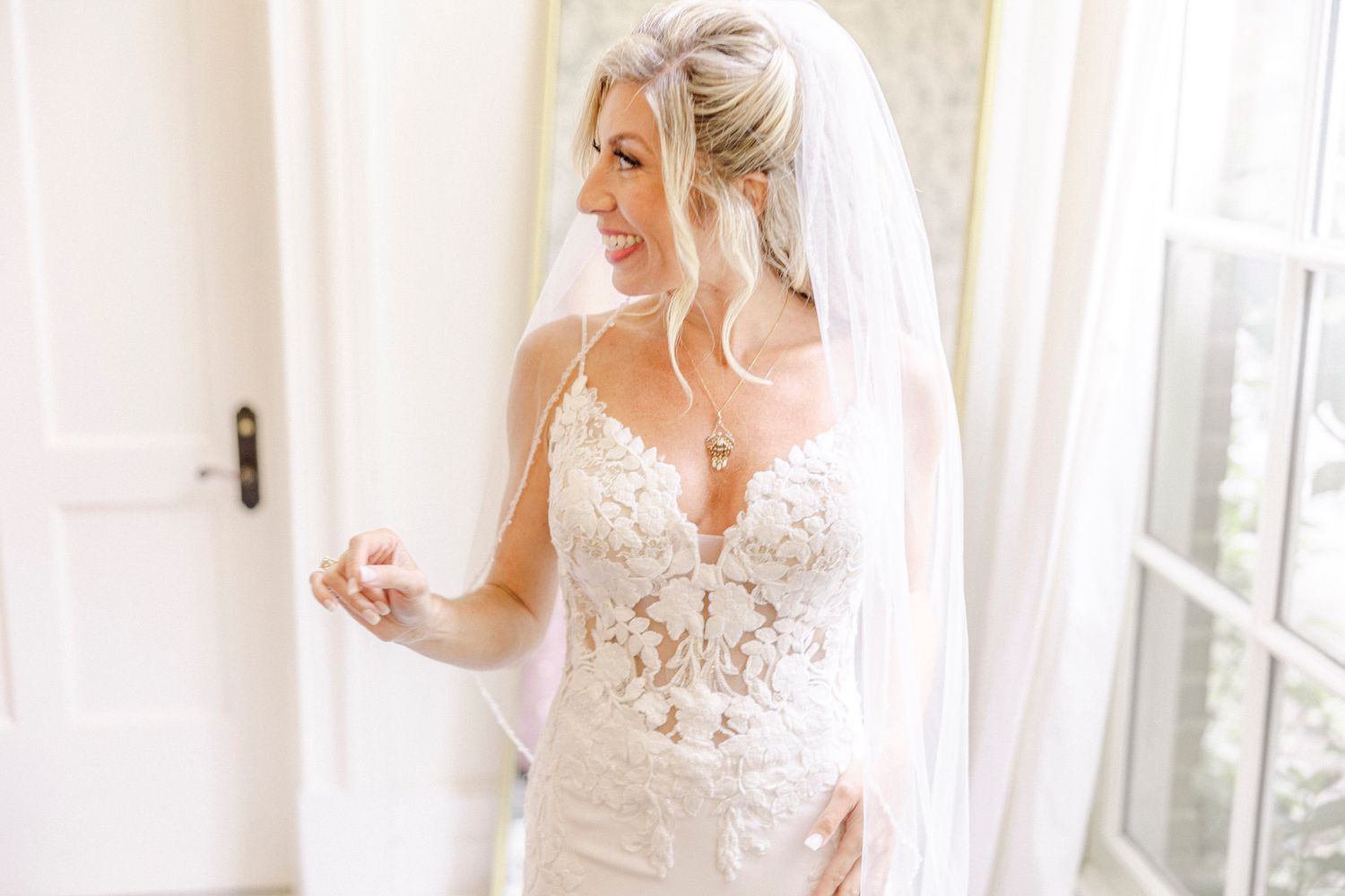 A bride in a white lace wedding dress adjusts her veil while getting ready in a sunlit room.