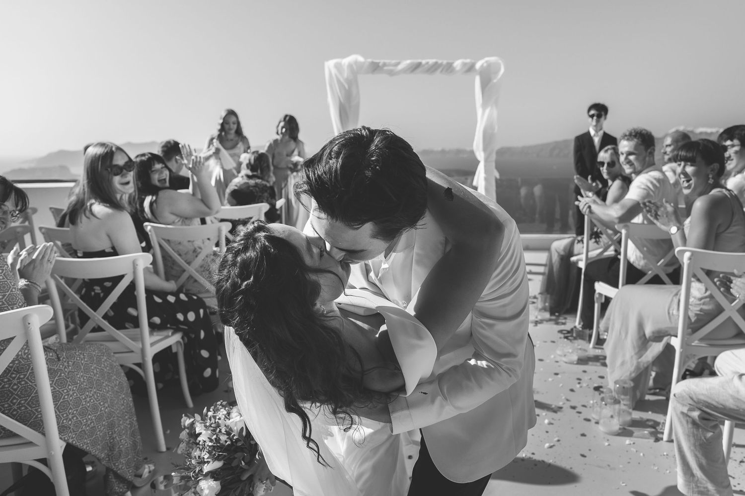 Black and white photo of wedding couple kissing at destination wedding ceremony with guests in the background applauding.
