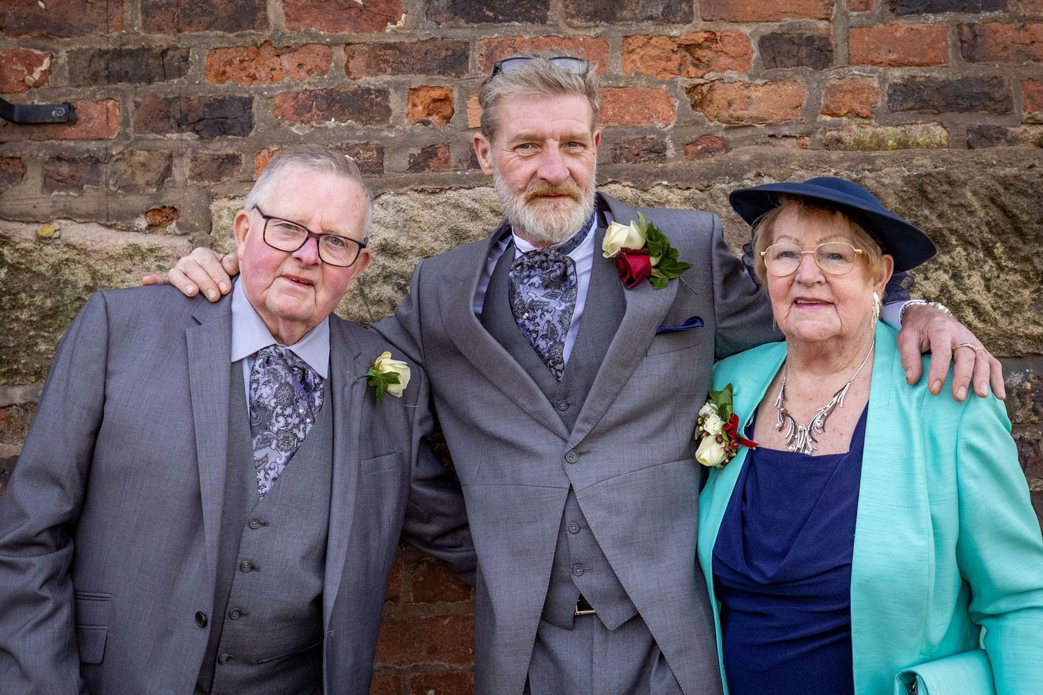 Groom with his parents against a brick wall at Windmill Farm, Burscough during relaxed family portraits after the Southport Town Hall weddin