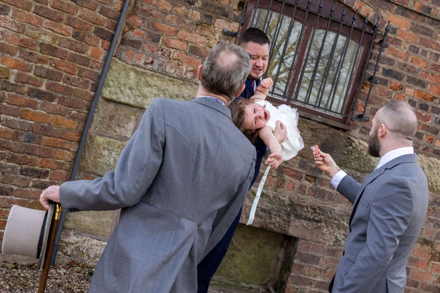 Groom & Groomsan laughing as a young flower girl protests during relaxed family photos outside the brick wall at Windmill Farm, Burscough.