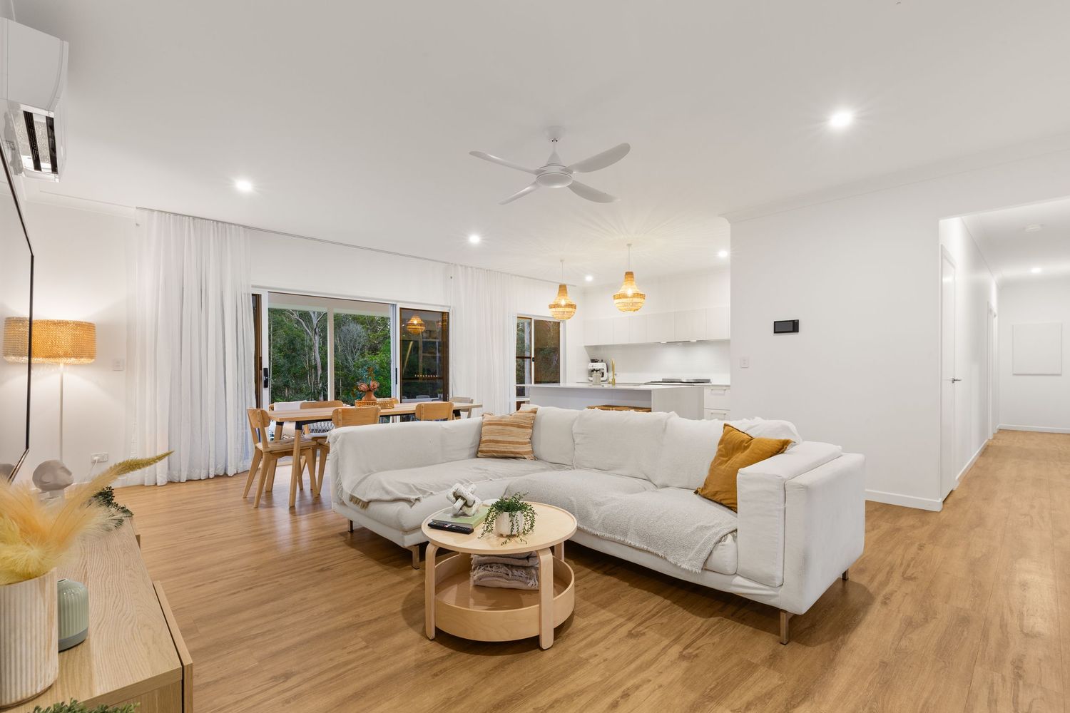 Bright open-plan living room with timber floors, white sofa, ceiling fan, and view to kitchen.