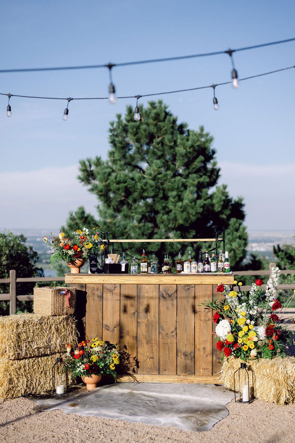 A rustic outdoor venue setup with wooden fencing, hay bales, and colorful floral arrangements under string lights.