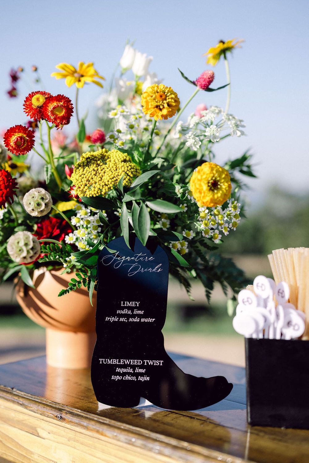 Colorful floral arrangements in black and gold vases displayed on wooden tables at a rustic outdoor event.