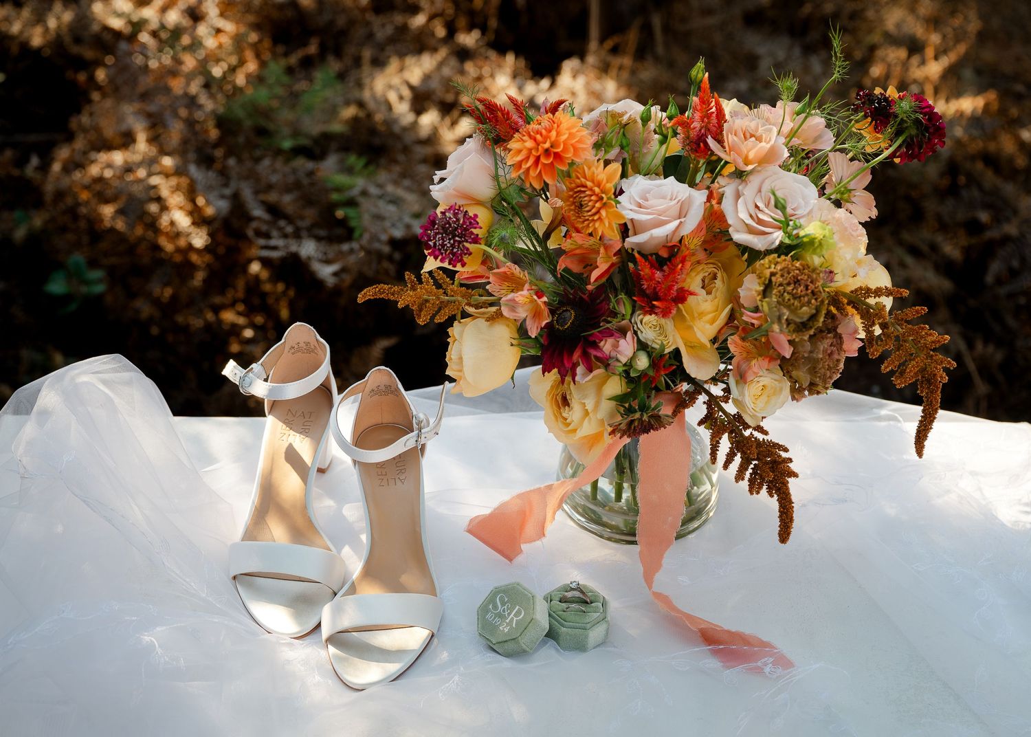 Wedding details with white bridal shoes and colorful fall floral arrangement in glass vase on white tablecloth.