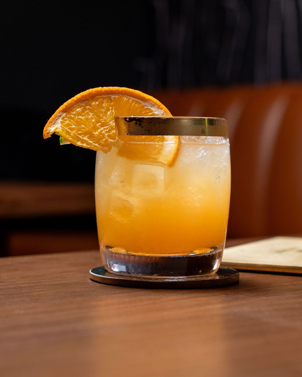 A bright orange cocktail with a citrus garnish served in a coupe glass on a dark wooden surface in dramatic lighting.