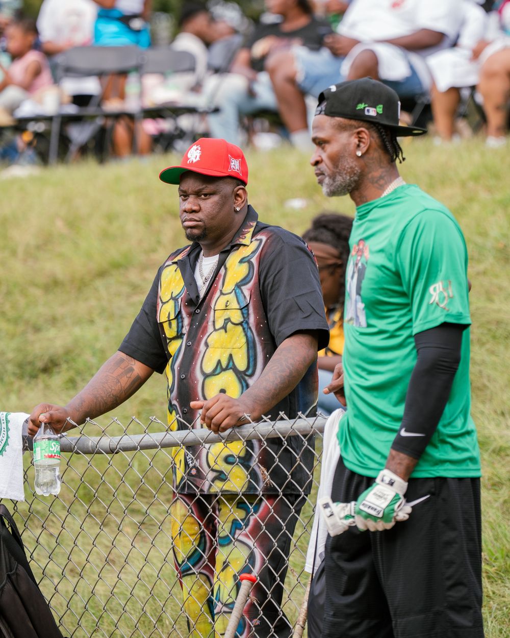 Two people stand by a chain link fence at an outdoor event, one wearing a colorful patterned shirt and the other in green.