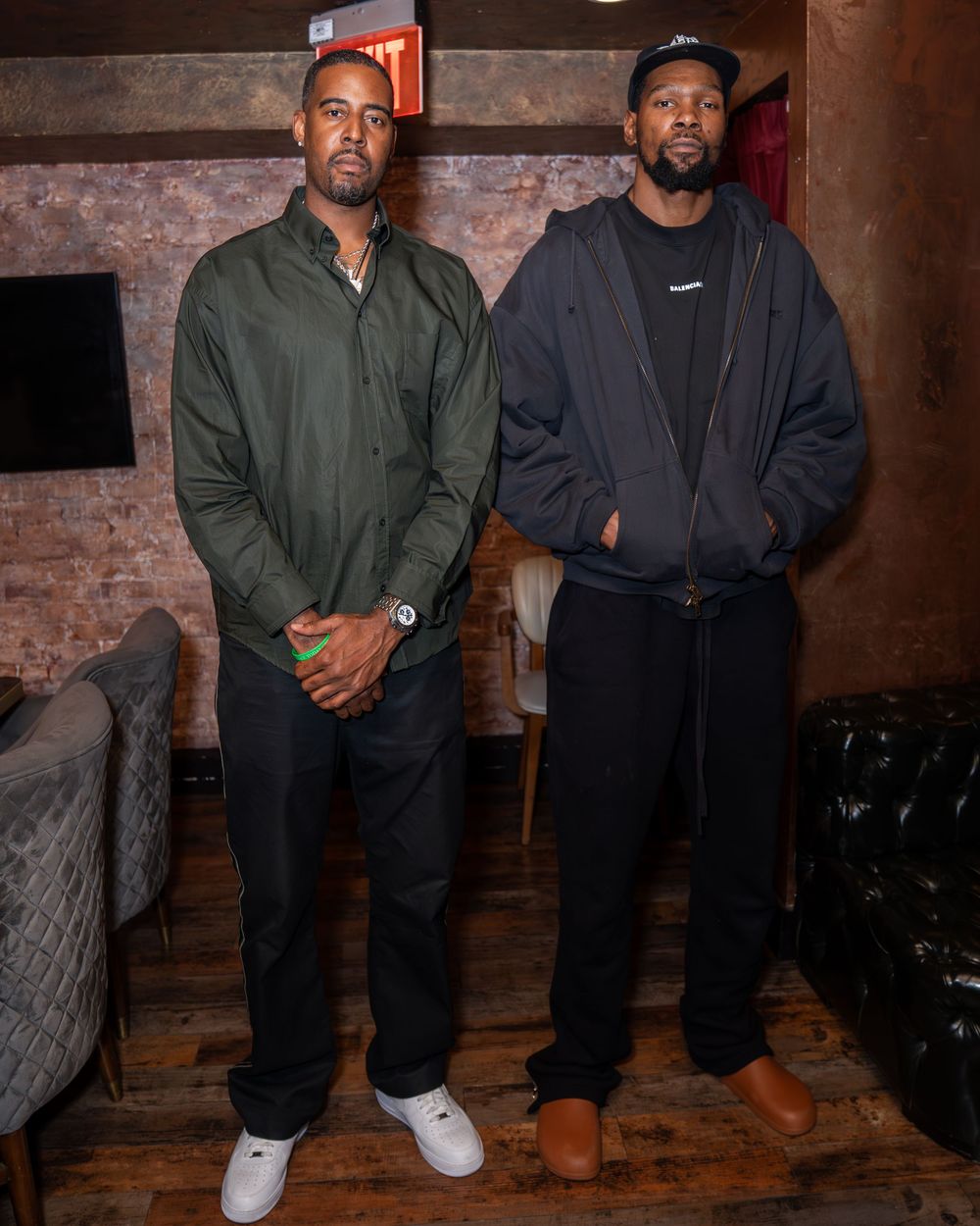 Two people in casual outfits standing together in front of a rustic brick wall in a dimly lit space.