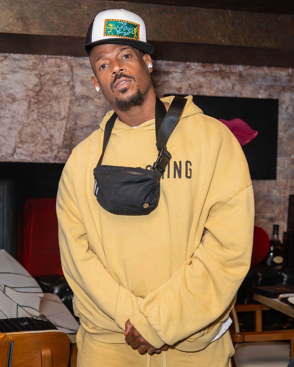 Person in a yellow sweatshirt and baseball cap poses in what appears to be a dimly lit indoor setting.