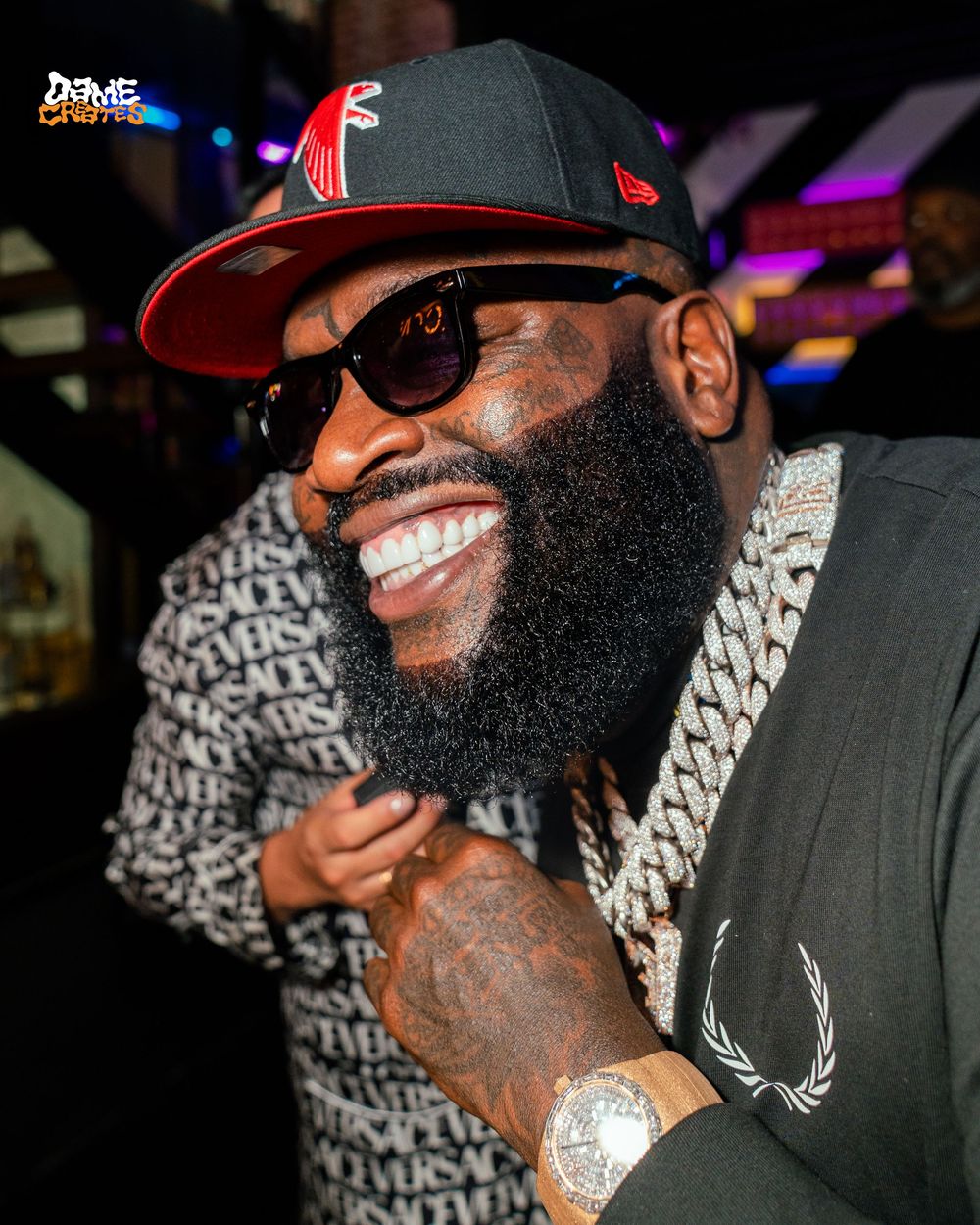 A bearded person wearing sunglasses and a thick silver chain necklace smiles while posing at an event with colorful lighting.