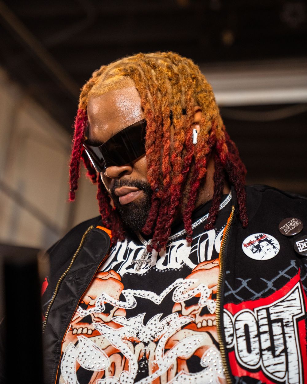 Professional wrestler wearing black and white graphic print jacket and orange-red dreadlocks performs in the ring.