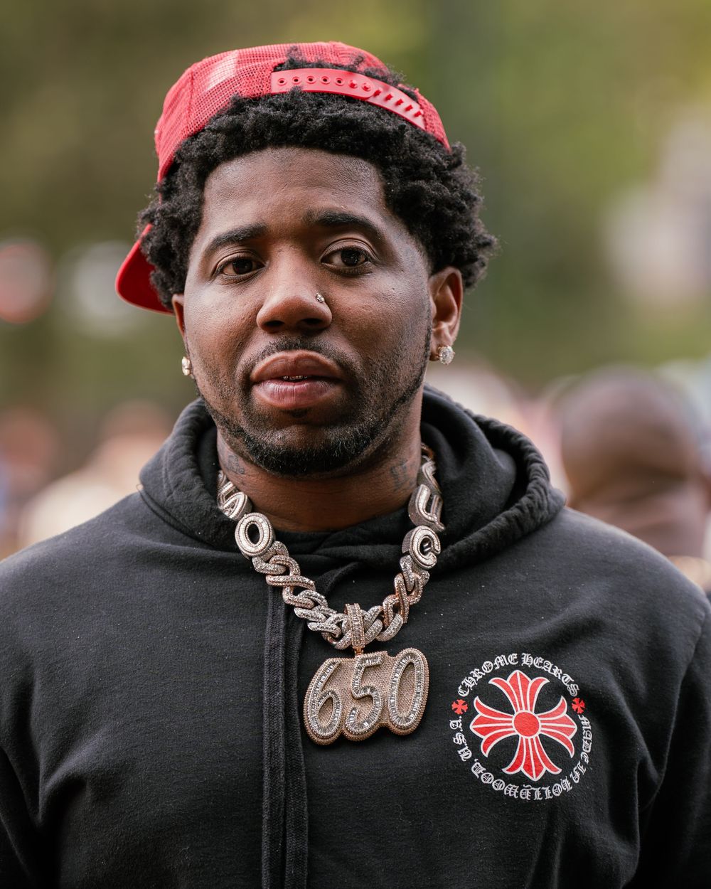 YFN LUCCI wearing a black hoodie and silver chain necklace with cross pendant looking at camera.