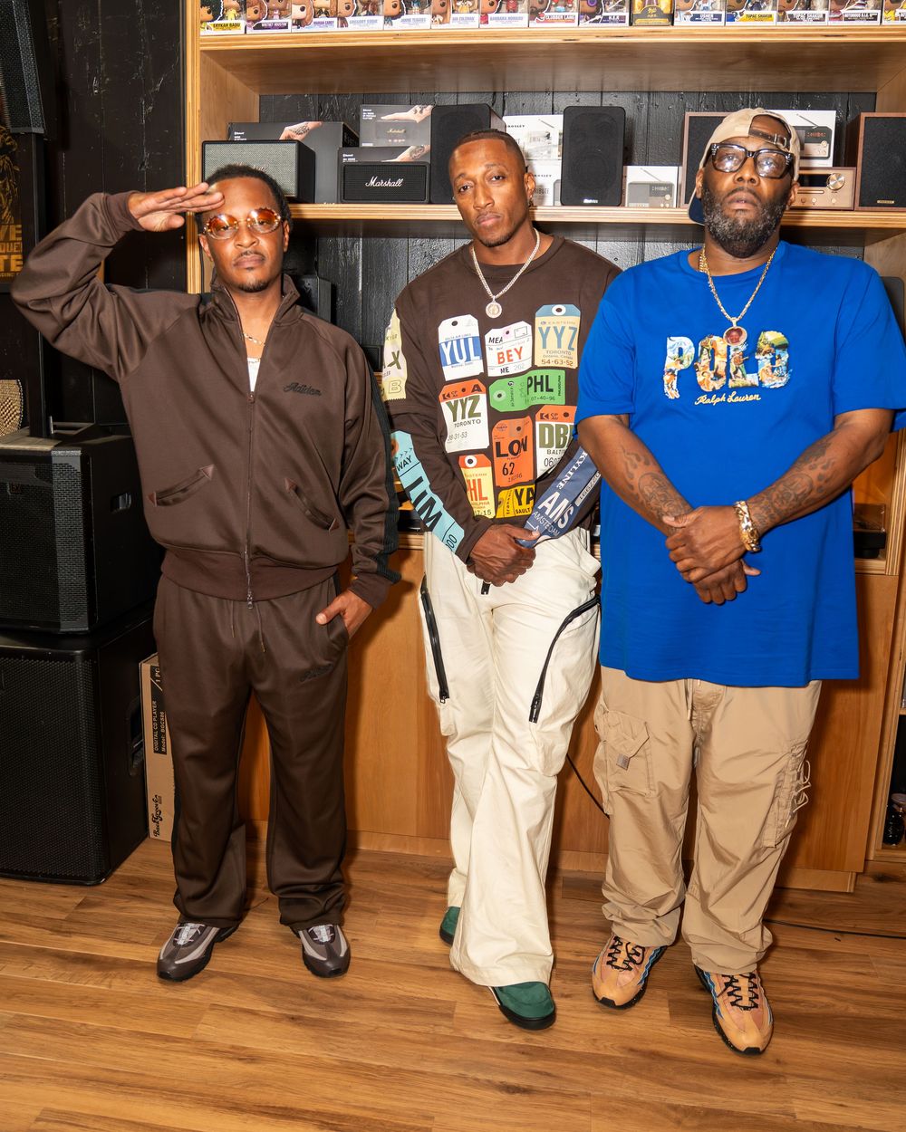 Three people pose together in a recording studio wearing casual streetwear including a blue polo shirt and brown jacket.