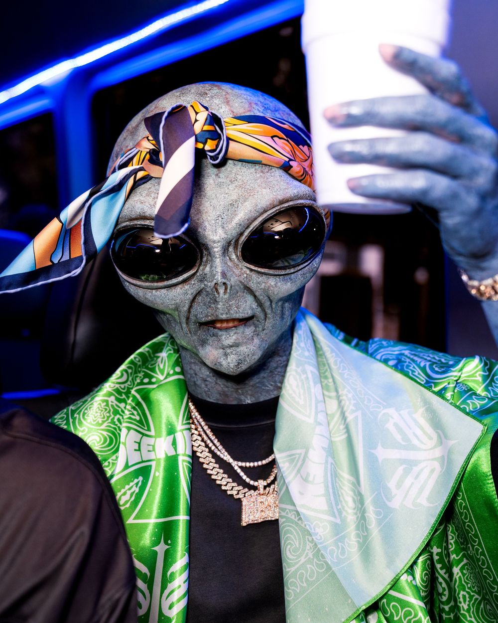 Person in alien mask wearing green shiny jacket and sunglasses at nightclub with blue lighting.