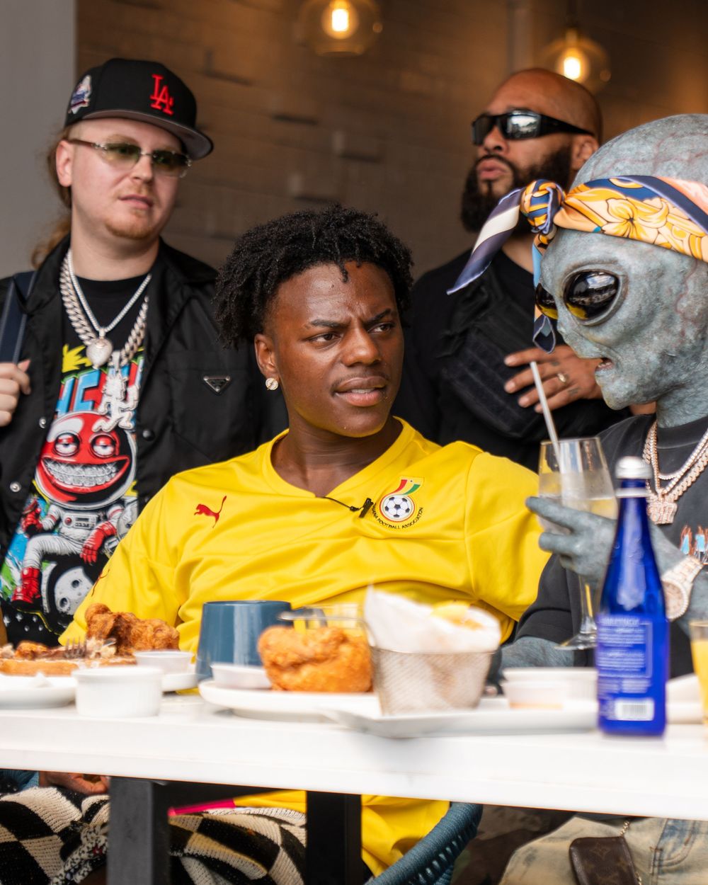 A group of people enjoying food and drinks at a restaurant table with yellow attire and baseball caps visible.