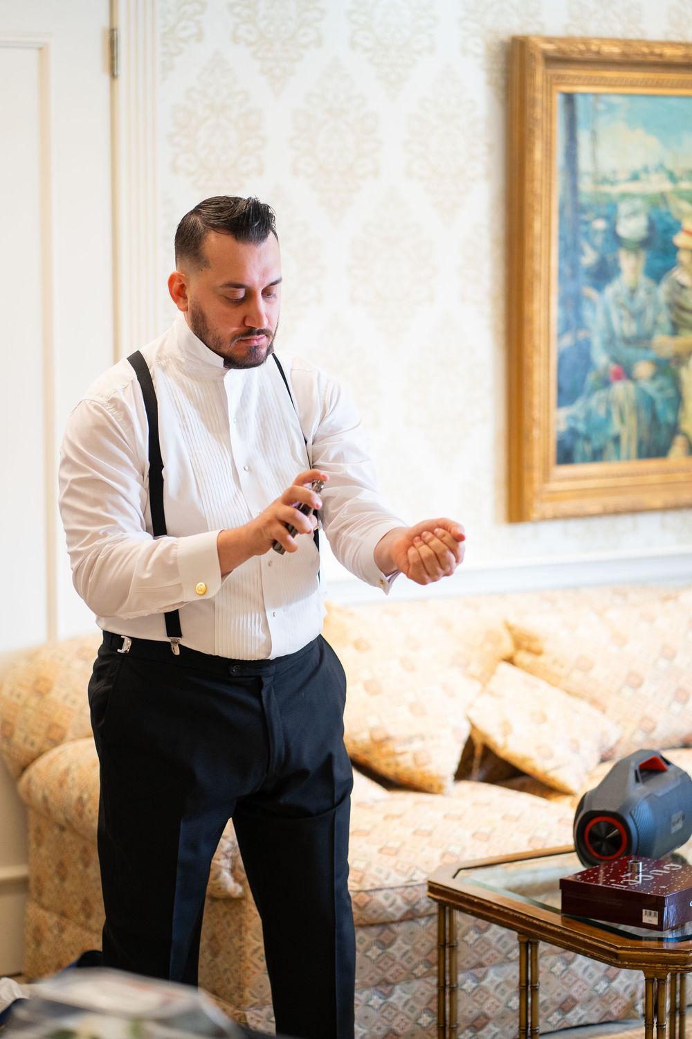 A groom in a white dress shirt and suspenders prepares for his wedding day in an elegant room with vintage decor.